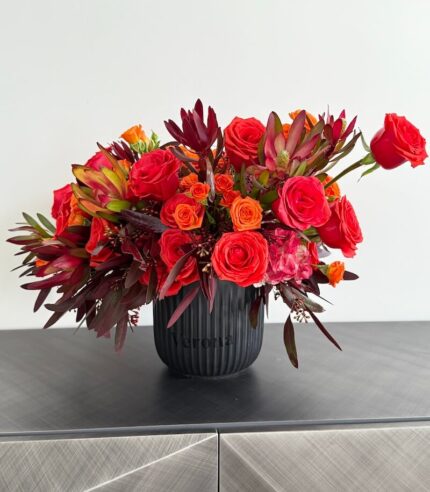 Lava Kiss bouquet with rich red roses, bright orange spray roses, exotic protea, and burgundy foliage in a sleek black ceramic vase – bold and dramatic.