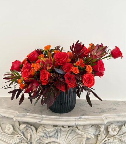 Lava Kiss bouquet with rich red roses, bright orange spray roses, exotic protea, and burgundy foliage in a sleek black ceramic vase – bold and dramatic.