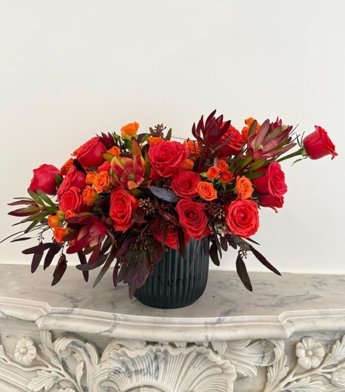 Lava Kiss bouquet with rich red roses, bright orange spray roses, exotic protea, and burgundy foliage in a sleek black ceramic vase – bold and dramatic.